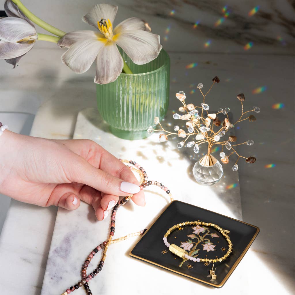 A hand holds a beaded necklace over marble next to the Jewelry Dish & Stud Earring Set - Flower/Black, gold jewelry, a glass ornament, and a green vase with a white flower. Sunlight casts rainbow reflections on the scene.
